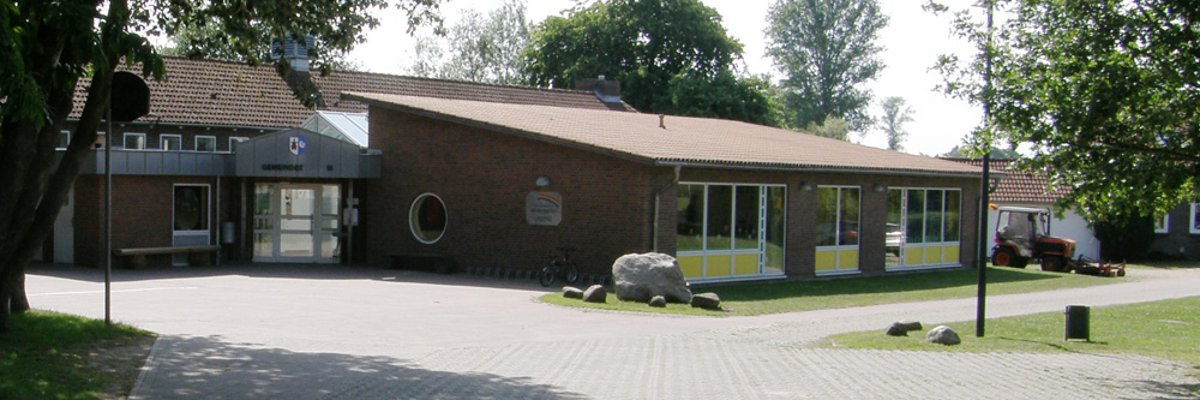 Backsteinbau des Gemeindezemtrums und Kindergartens von Labenz mit Eingangsbereich, großen Fenstern und einem gepflasterten Vorplatz, umgeben von Bäumen und Grünflächen.