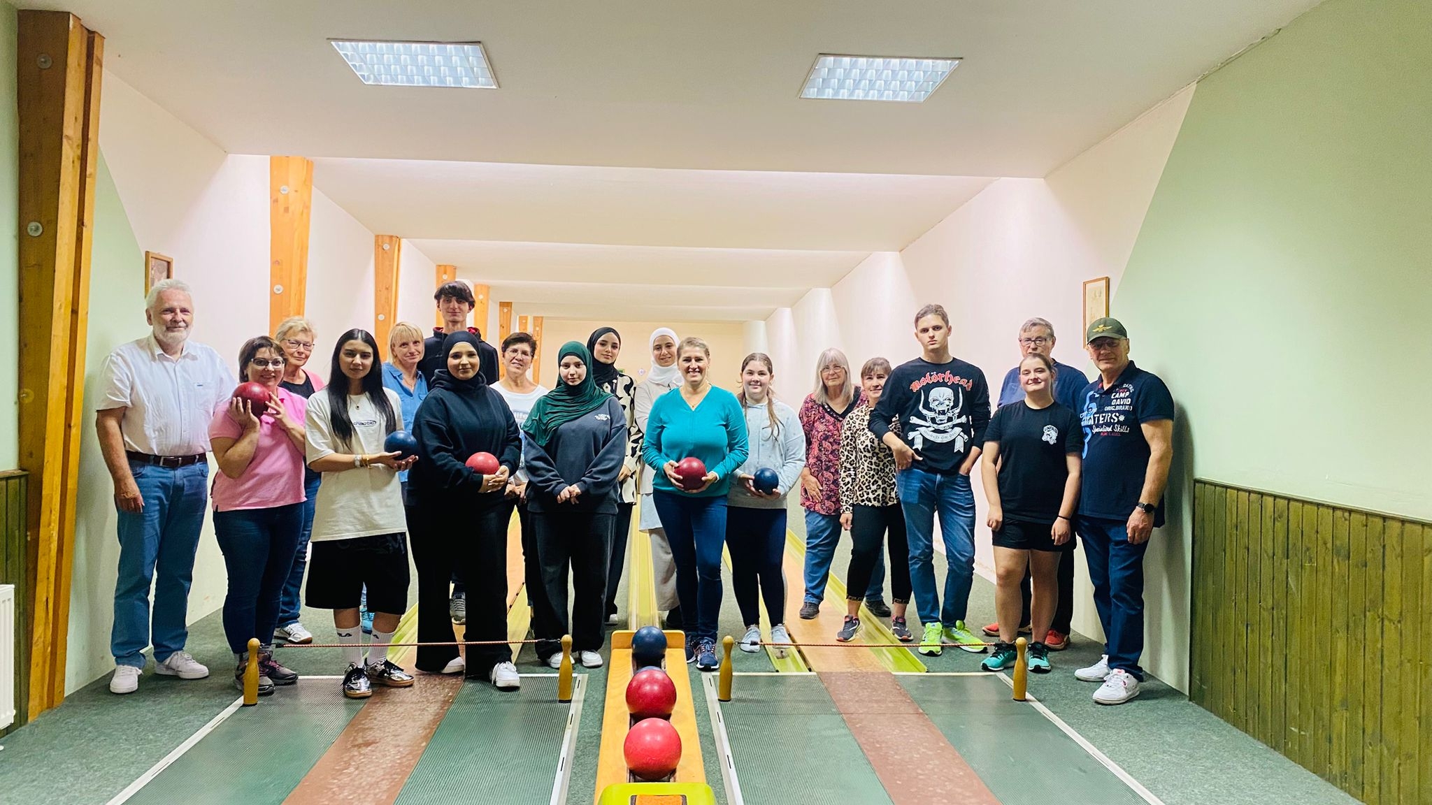 Das Bild zeigt eine Gruppe von Personen, die auf einer Kegelbahn posieren. Die Gruppe besteht aus Menschen verschiedenen Alters, die Kegelkugeln in den Händen halten. Sie stehen in zwei Reihen nebeneinander vor den Kegelbahnen. Im Hintergrund sind die Kegel zu sehen. Der Raum ist beleuchtet, und die Wände sind in hellen Farben gehalten.