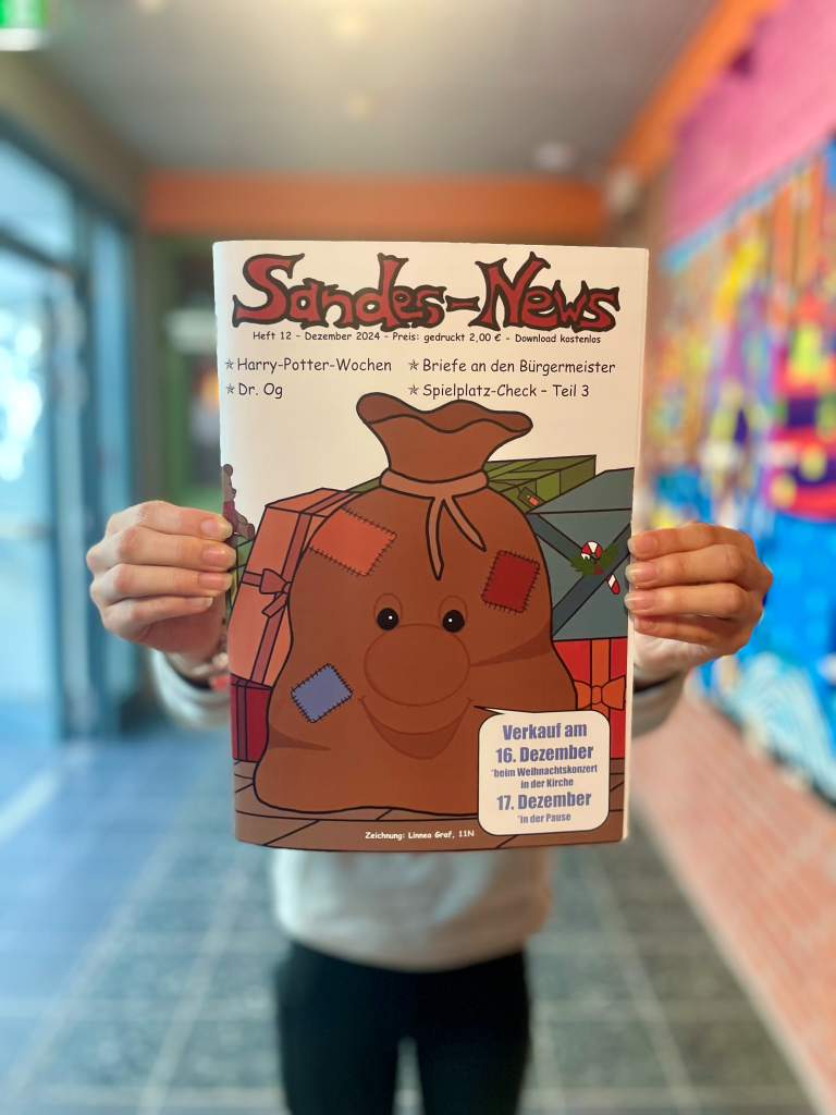 Eine Person hält die Ausgabe 12 (Dezember 2024) der Schülerzeitung „Sandes-News“ in die Kamera. Auf dem Titel ist ein fröhlich lächelnder Geschenkesack mit bunten Flicken gezeichnet, im Hintergrund liegen Geschenke. Themen der Ausgabe sind u. a. „Harry-Potter-Wochen“, „Dr. Og“, „Briefe an den Bürgermeister“ und „Spielplatz-Check – Teil 3“.