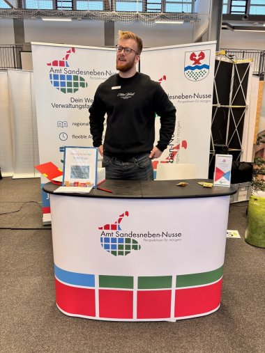 Das Bild zeigt einen Messe-Stand des Amtes Sandesneben-Nusse in einer Sporthallen-Umgebung. Hinter dem Stand stehen zwei große Roll-Up-Banner mit dem Logo und Schriftzug des Amtes. Vor dem Banner steht ein Mann mit Brille und dunklem Pullover hinter einem gebogenen Messtresen, der ebenfalls mit dem Logo des Amtes gestaltet ist.