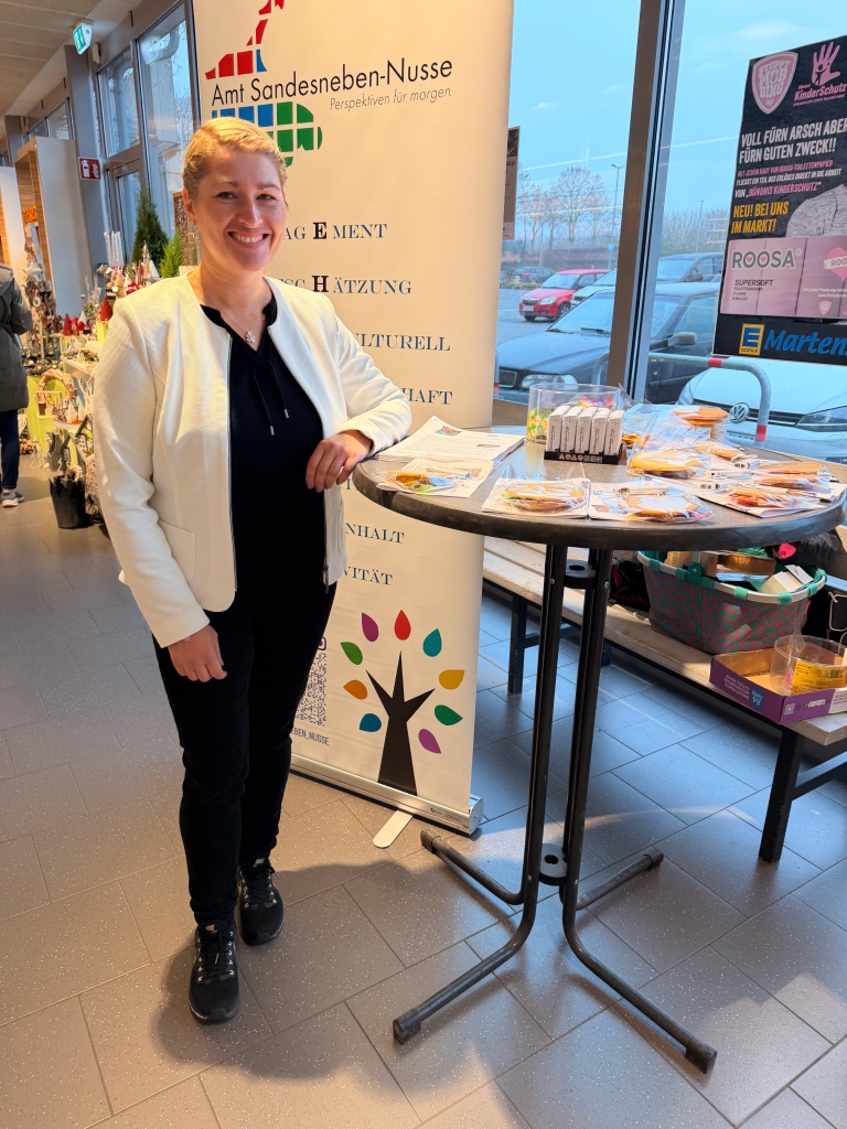 Eine Mitarbeiterin des Amtes Sandesneben-Nusse steht lächelnd neben einem Roll-Up-Banner und einem Stehtisch mit Informationsmaterial in einem Eingangsbereich.