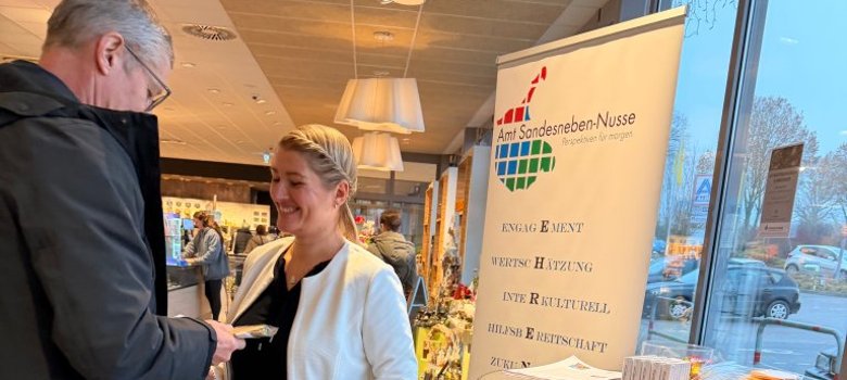 Eine Mitarbeiterin des Amtes Sandesneben-Nusse übergibt einem Besucher Informationsmaterial an einem Stehtisch; daneben steht ein Roll-Up-Banner des Amtes im Hintergrund.