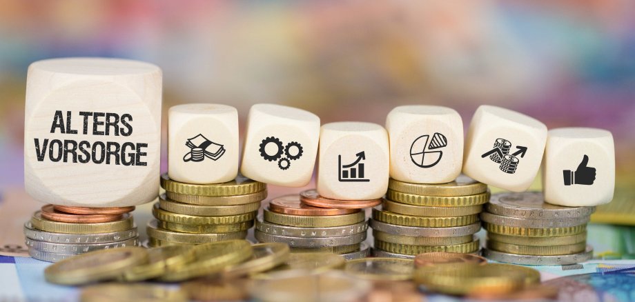 Symbolfoto zur Altersvorsorge: Aufgestapelte Münzen mit Holz­würfeln, auf denen Begriffe und Symbole zu Finanzen, Wachstum und Investitionen abgebildet sind. Auf dem größten Würfel steht das Wort „Altersvorsorge“.