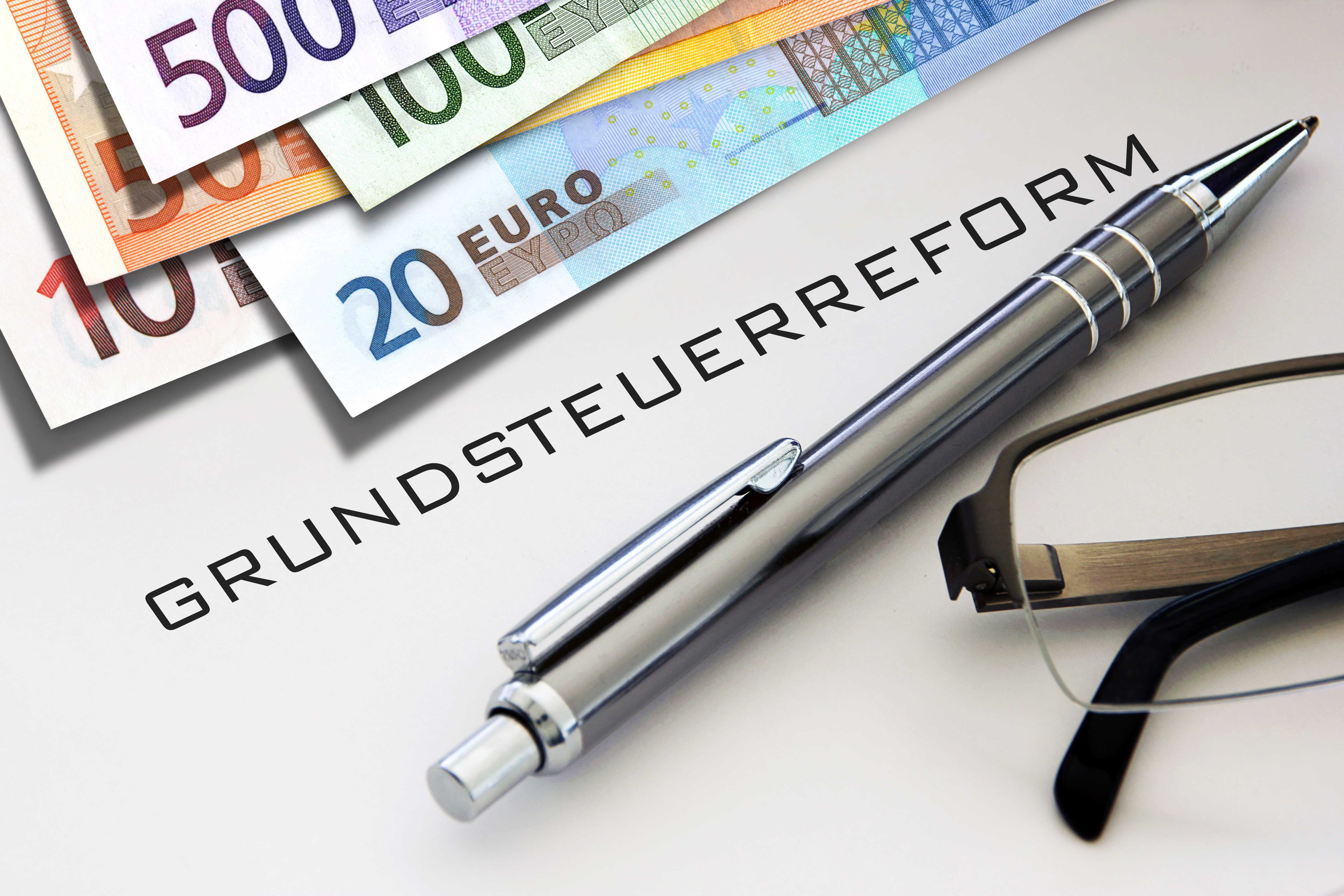 Symbolfoto zur Grundsteuerreform: Auf einem Schreibtisch liegen Euro-Banknoten, ein Kugelschreiber und eine Brille. In der Mitte steht das Wort „Grundsteuerreform“.