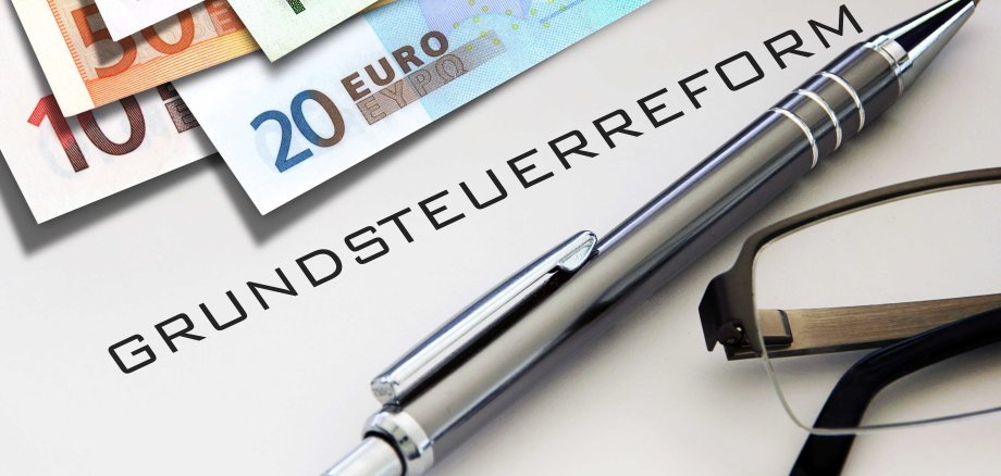 Symbolfoto zur Grundsteuerreform: Auf einem Schreibtisch liegen Euro-Banknoten, ein Kugelschreiber und eine Brille. In der Mitte steht das Wort „Grundsteuerreform“.