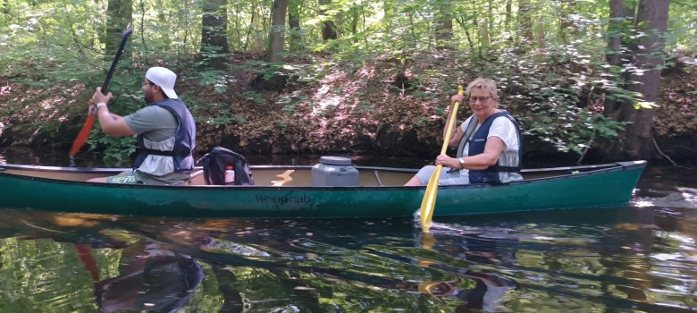 Zwei Personen paddeln gemeinsam in einem grünen Kanu durch einen von Bäumen gesäumten Fluss.