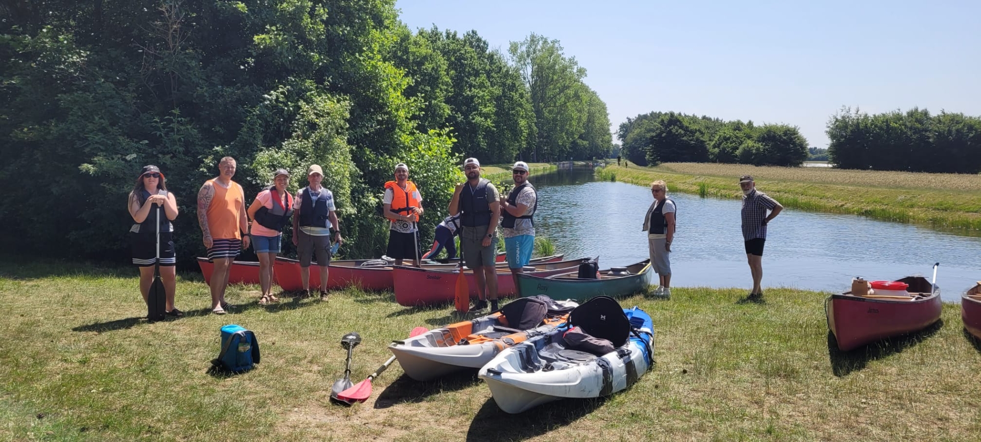 Eine Gruppe von Teilnehmenden steht mit Paddeln und Kanus am Flussufer, bereit zur Kanutour bei sonnigem Wetter.