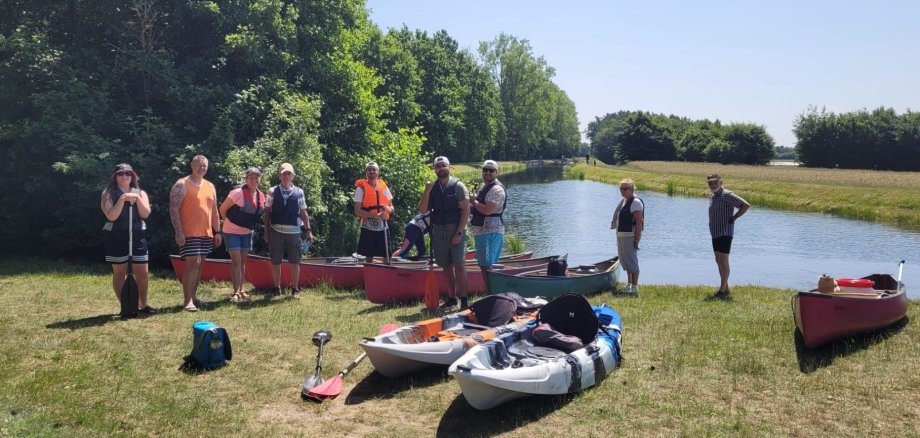 Eine Gruppe von Teilnehmenden steht mit Paddeln und Kanus am Flussufer, bereit zur Kanutour bei sonnigem Wetter.