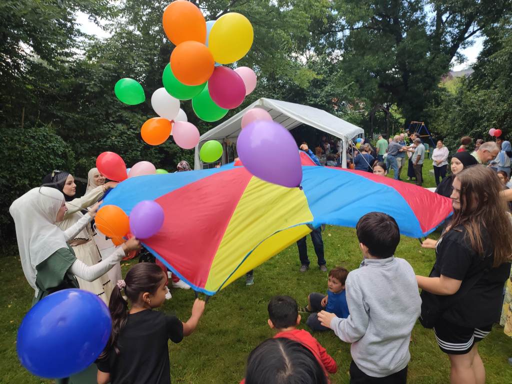 Kinder und Erwachsene spielen beim Sommerfest mit einem bunten Schwungtuch und vielen Luftballons.