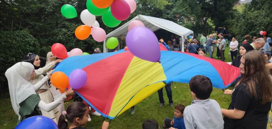 Kinder und Erwachsene spielen beim Sommerfest mit einem bunten Schwungtuch und vielen Luftballons.