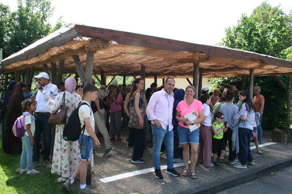 Eine Gruppe von Menschen – darunter Frauen, Männer und Kinder – steht unter einem offenen, schattenspendenden Holzunterstand mit Netzabdeckung im Serengeti-Park. Im Vordergrund stehen ein Mann und eine Frau und lächeln in die Kamera, während sich hinter ihnen viele Teilnehmende unterhalten oder auf Pläne und Infoblätter schauen. Die Szene wirkt lebendig und erwartungsvoll – ein Moment zu Beginn oder in einer Pause des gemeinsamen Ausflugstages.