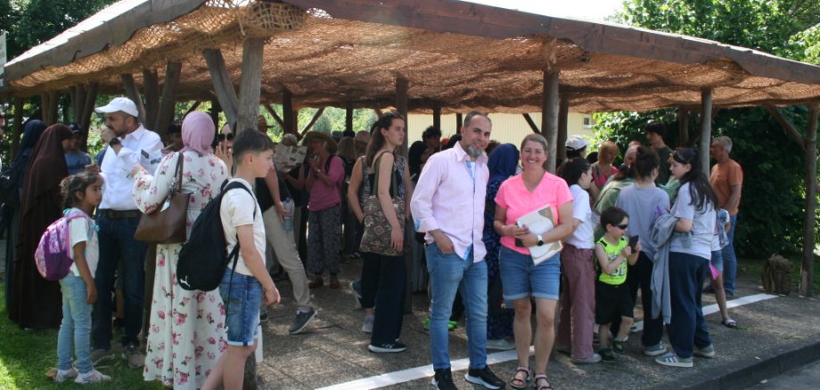 Eine Gruppe von Menschen – darunter Frauen, Männer und Kinder – steht unter einem offenen, schattenspendenden Holzunterstand mit Netzabdeckung im Serengeti-Park. Im Vordergrund stehen ein Mann und eine Frau und lächeln in die Kamera, während sich hinter ihnen viele Teilnehmende unterhalten oder auf Pläne und Infoblätter schauen. Die Szene wirkt lebendig und erwartungsvoll – ein Moment zu Beginn oder in einer Pause des gemeinsamen Ausflugstages.