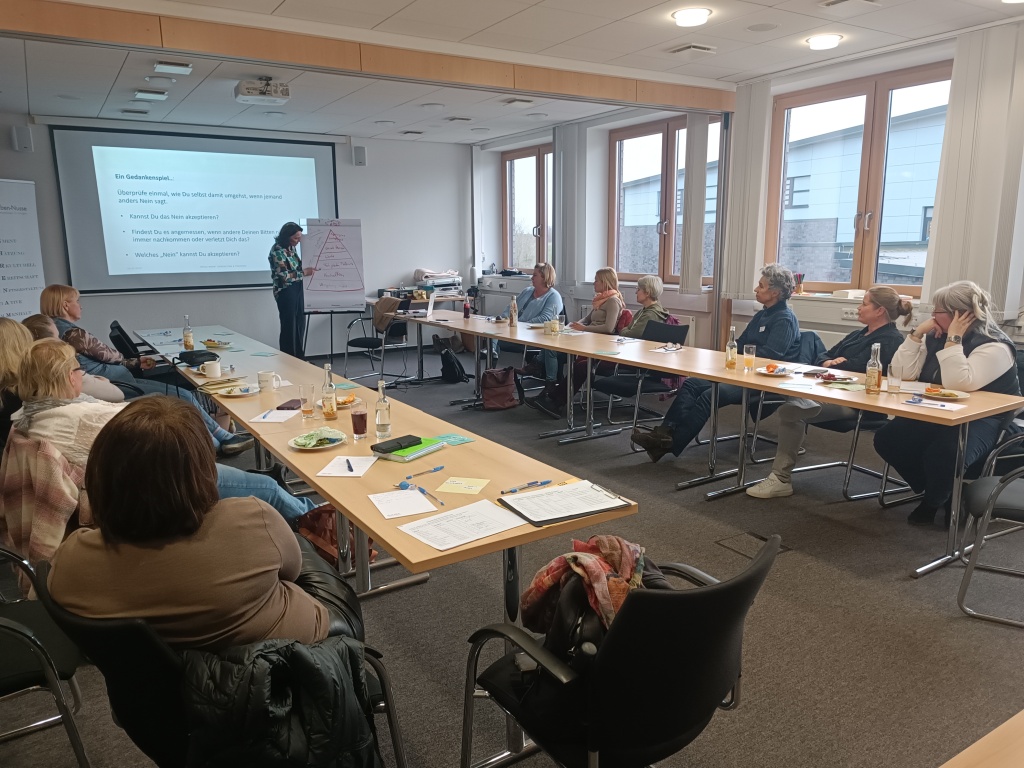 Teilnehmende des Workshops „Gehört werden im Ehrenamt – Die Kunst des Nein-Sagens und Grenzen-Setzens“ sitzen in einem modernen Seminarraum an einem U-förmig angeordneten Tisch. Eine Referentin steht vorne und erklärt mithilfe einer Präsentationsfolie und einem Flipchart, auf dem eine Pyramide skizziert ist. Auf der Leinwand ist ein Gedankenexperiment zum Thema Nein-Sagen zu sehen. Die Teilnehmenden hören aufmerksam zu, machen sich Notizen und haben Getränke sowie kleine Snacks vor sich. Die Atmosphäre ist konzentriert und konstruktiv.