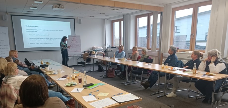 Teilnehmende des Workshops „Gehört werden im Ehrenamt – Die Kunst des Nein-Sagens und Grenzen-Setzens“ sitzen in einem modernen Seminarraum an einem U-förmig angeordneten Tisch. Eine Referentin steht vorne und erklärt mithilfe einer Präsentationsfolie und einem Flipchart, auf dem eine Pyramide skizziert ist. Auf der Leinwand ist ein Gedankenexperiment zum Thema Nein-Sagen zu sehen. Die Teilnehmenden hören aufmerksam zu, machen sich Notizen und haben Getränke sowie kleine Snacks vor sich. Die Atmosphäre ist konzentriert und konstruktiv.