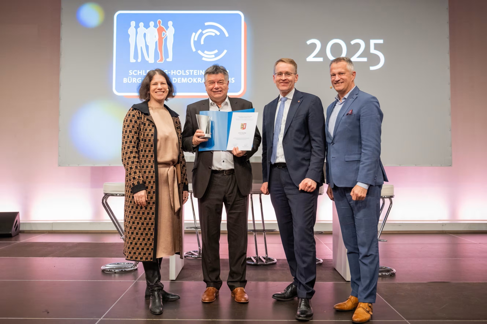 Ulrich Hardtke, Bürgermeister der Gemeinde Labenz, steht mit Kristina Herbst, Daniel Günther und Oliver Stolz bei der Verleihung des Demokratiepreises 2025 auf einer Bühne. Hardtke hält die Urkunde und die Auszeichnung in den Händen.