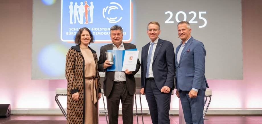 Ulrich Hardtke, Bürgermeister der Gemeinde Labenz, steht mit Kristina Herbst, Daniel Günther und Oliver Stolz bei der Verleihung des Demokratiepreises 2025 auf einer Bühne. Hardtke hält die Urkunde und die Auszeichnung in den Händen.