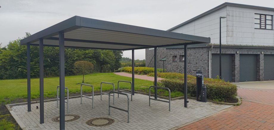 Überdachte Fahrradabstellanlage mit mehreren Fahrradständern und einer Fahrradreparaturstation vor einem modernen Gebäude.