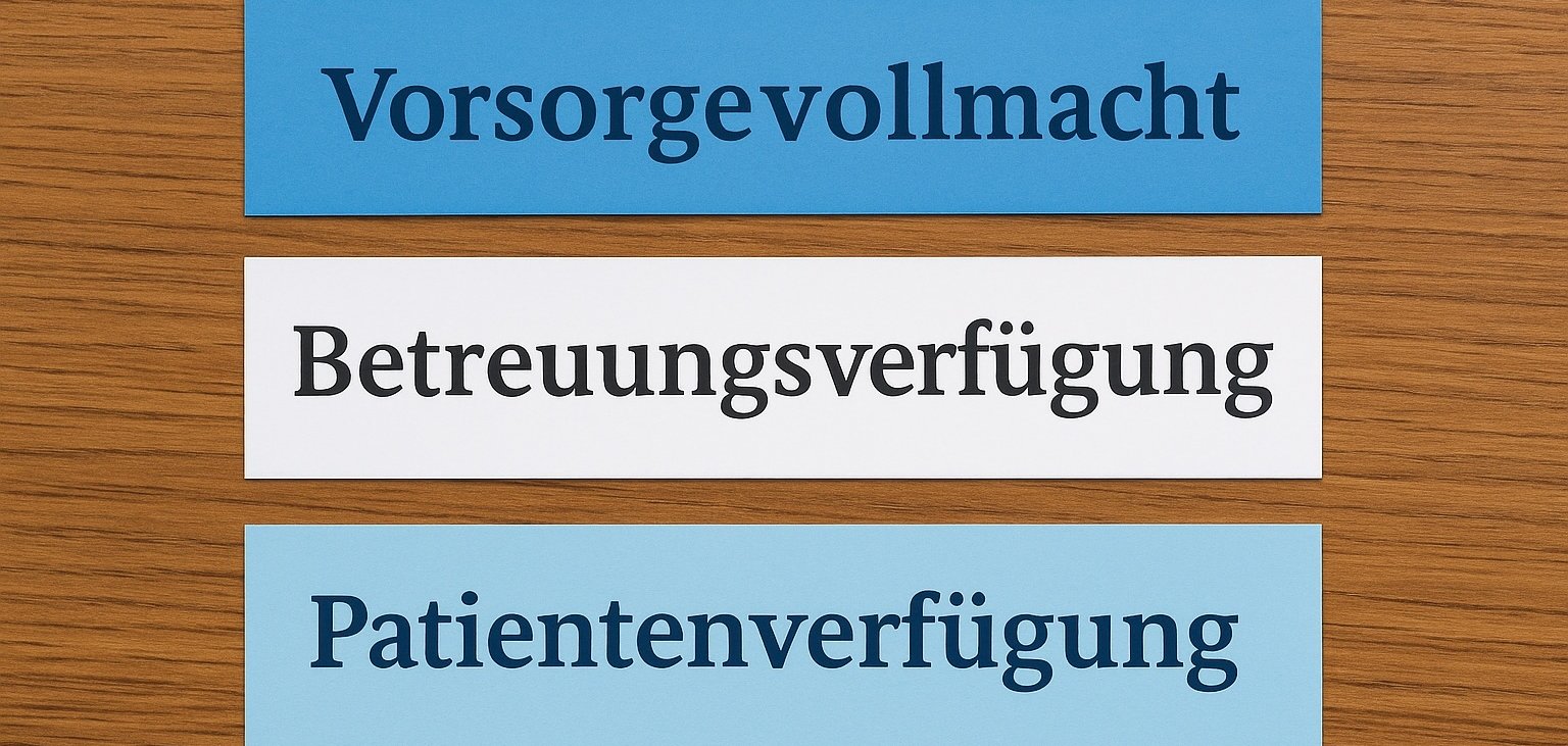 Drei farbige Papierstreifen mit den Aufschriften ‚Vorsorgevollmacht‘, ‚Betreuungsverfügung‘ und ‚Patientenverfügung‘ liegen nebeneinander auf einer Holzoberfläche. Die Streifen sind in Blau- und Weißtönen gehalten.