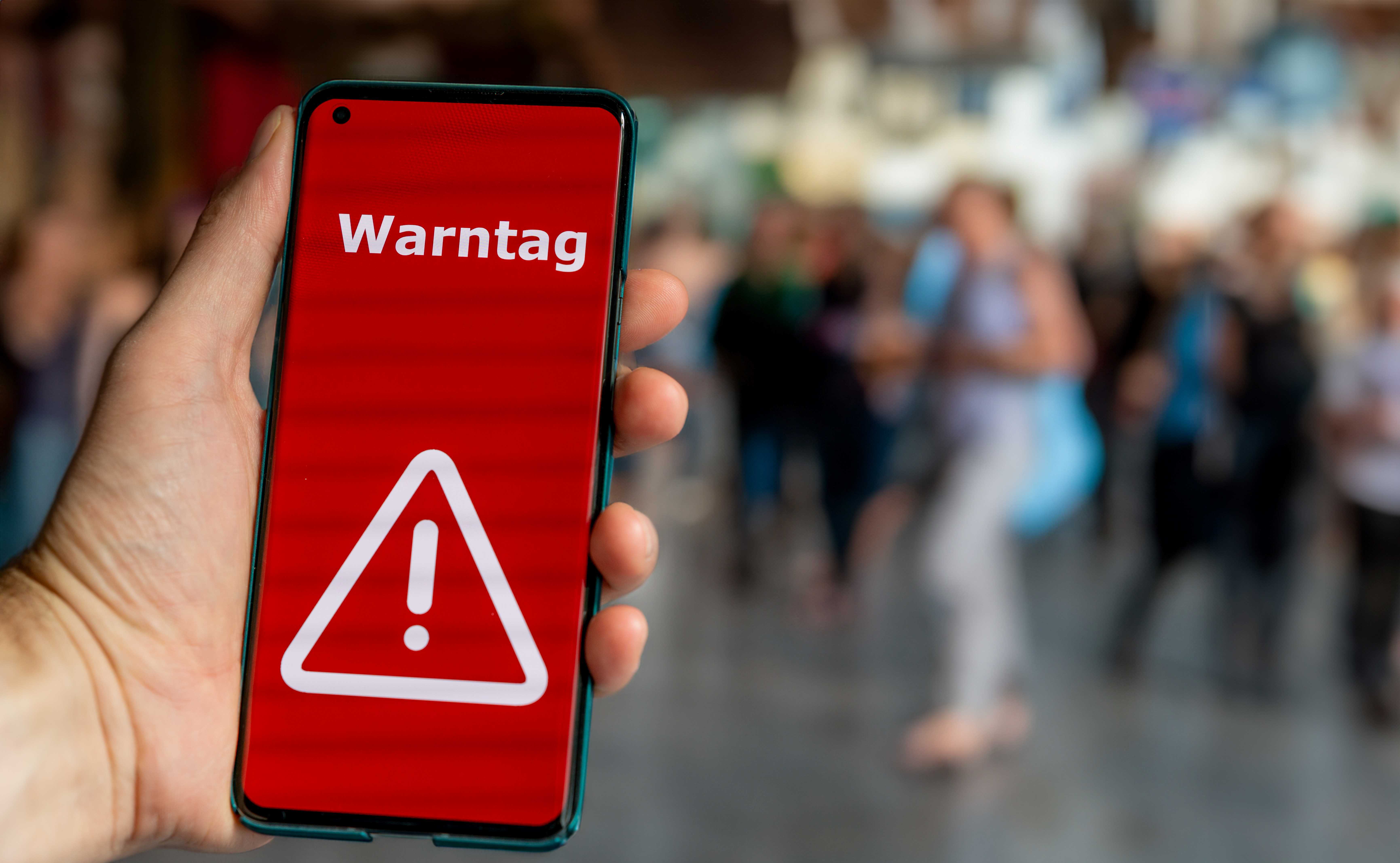 Hand hält ein Smartphone mit rotem Warnbildschirm und dem Schriftzug „Warntag“ in einer belebten Umgebung.