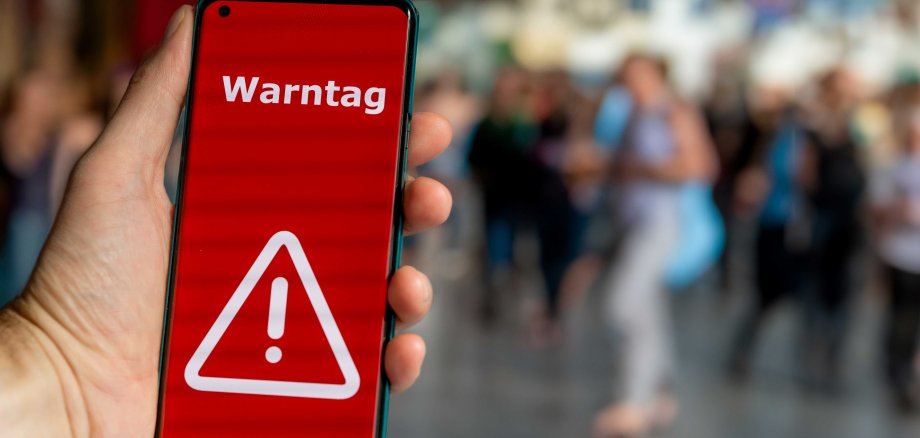 Hand hält ein Smartphone mit rotem Warnbildschirm und dem Schriftzug „Warntag“ in einer belebten Umgebung.