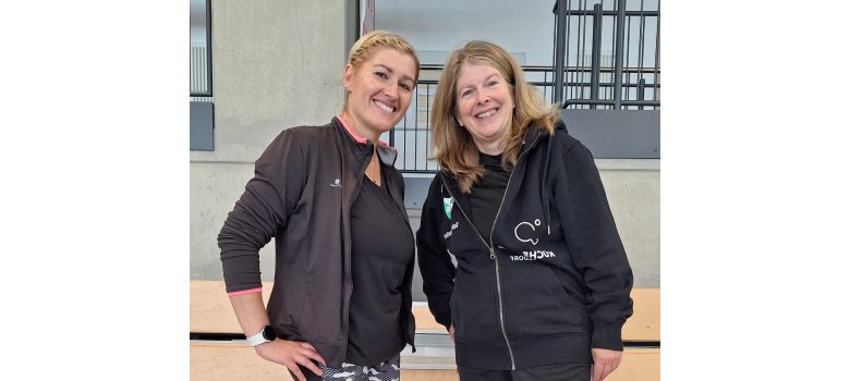Zwei Frauen in Sportkleidung lächeln in einer Sporthalle in die Kamera.