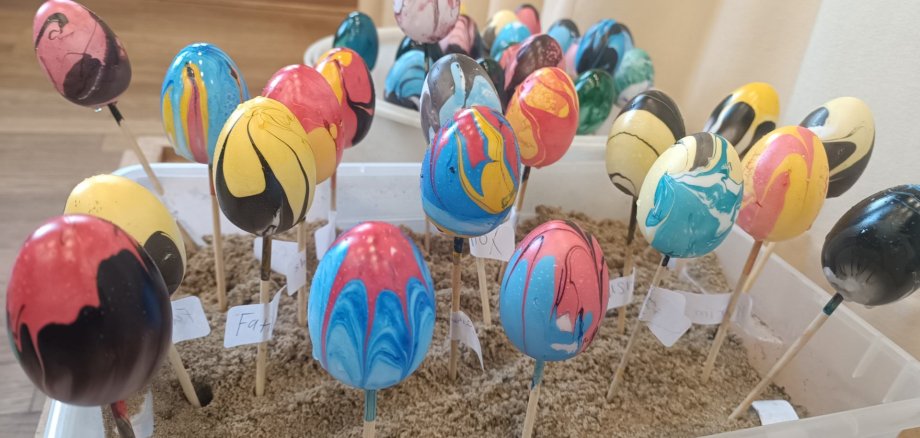 Bunt gefärbte Ostereier mit marmorierten Mustern stecken auf Holzstäbchen in einer Kiste mit Sand zum Trocknen.