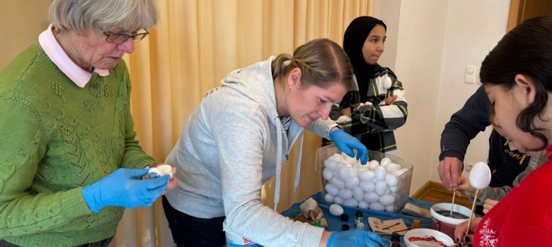 Mehrere Erwachsene und Kinder stehen an einem Tisch und färben gemeinsam Ostereier. Auf dem Tisch befinden sich Farbbecher, Handschuhe und Eier.
