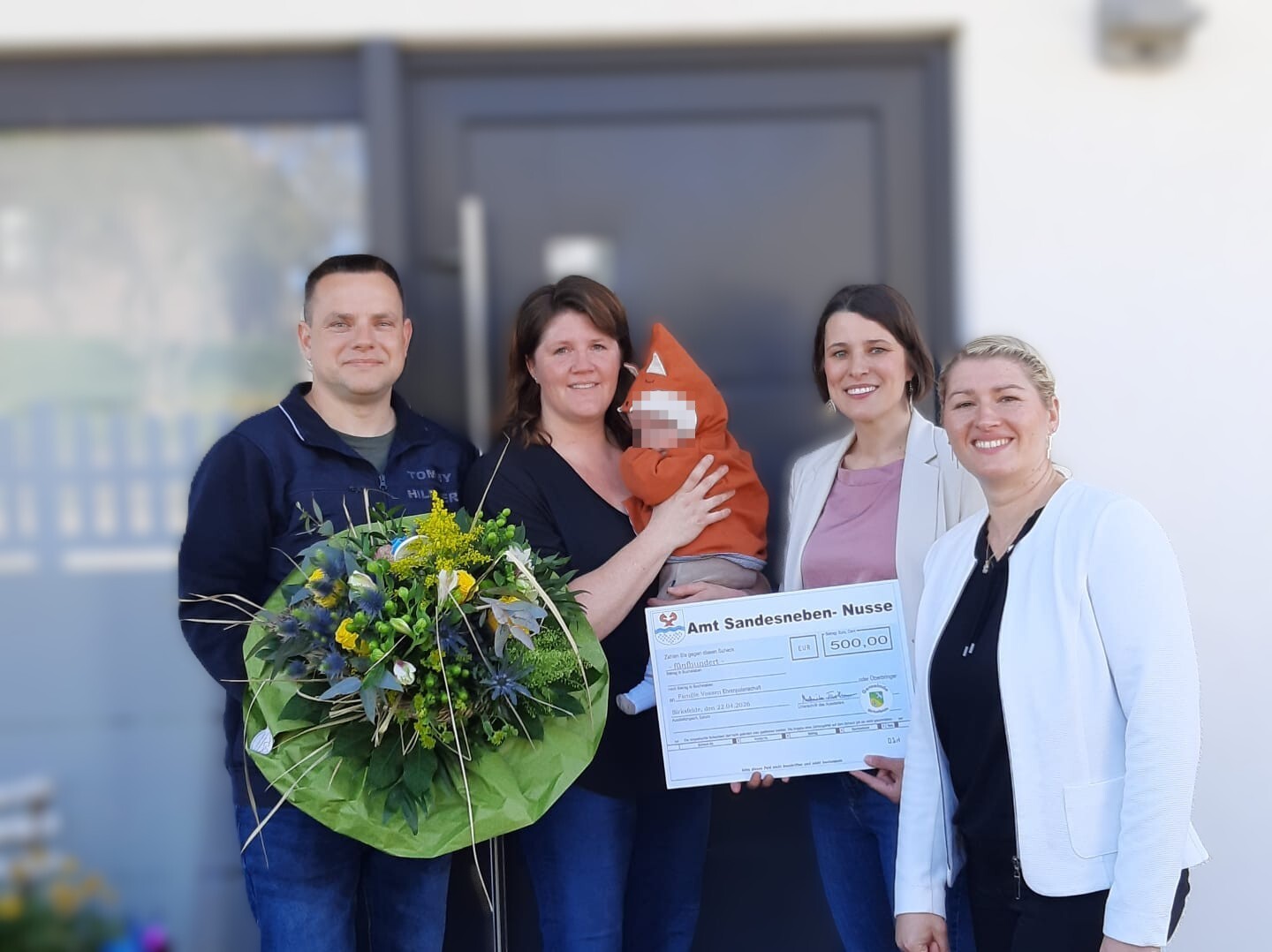 Fünf Personen stehen vor einem Gebäude und schauen in die Kamera. In der Mitte hält eine Frau ein Baby im Arm. Zwei Frauen rechts halten einen großen symbolischen Scheck über 500 Euro mit der Aufschrift „Amt Sandesneben-Nusse“. Links steht ein Mann mit einem großen Blumenstrauß. Alle Personen lächeln.