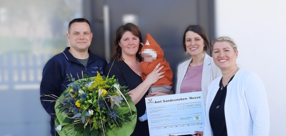 Fünf Personen stehen vor einem Gebäude und schauen in die Kamera. In der Mitte hält eine Frau ein Baby im Arm. Zwei Frauen rechts halten einen großen symbolischen Scheck über 500 Euro mit der Aufschrift „Amt Sandesneben-Nusse“. Links steht ein Mann mit einem großen Blumenstrauß. Alle Personen lächeln.