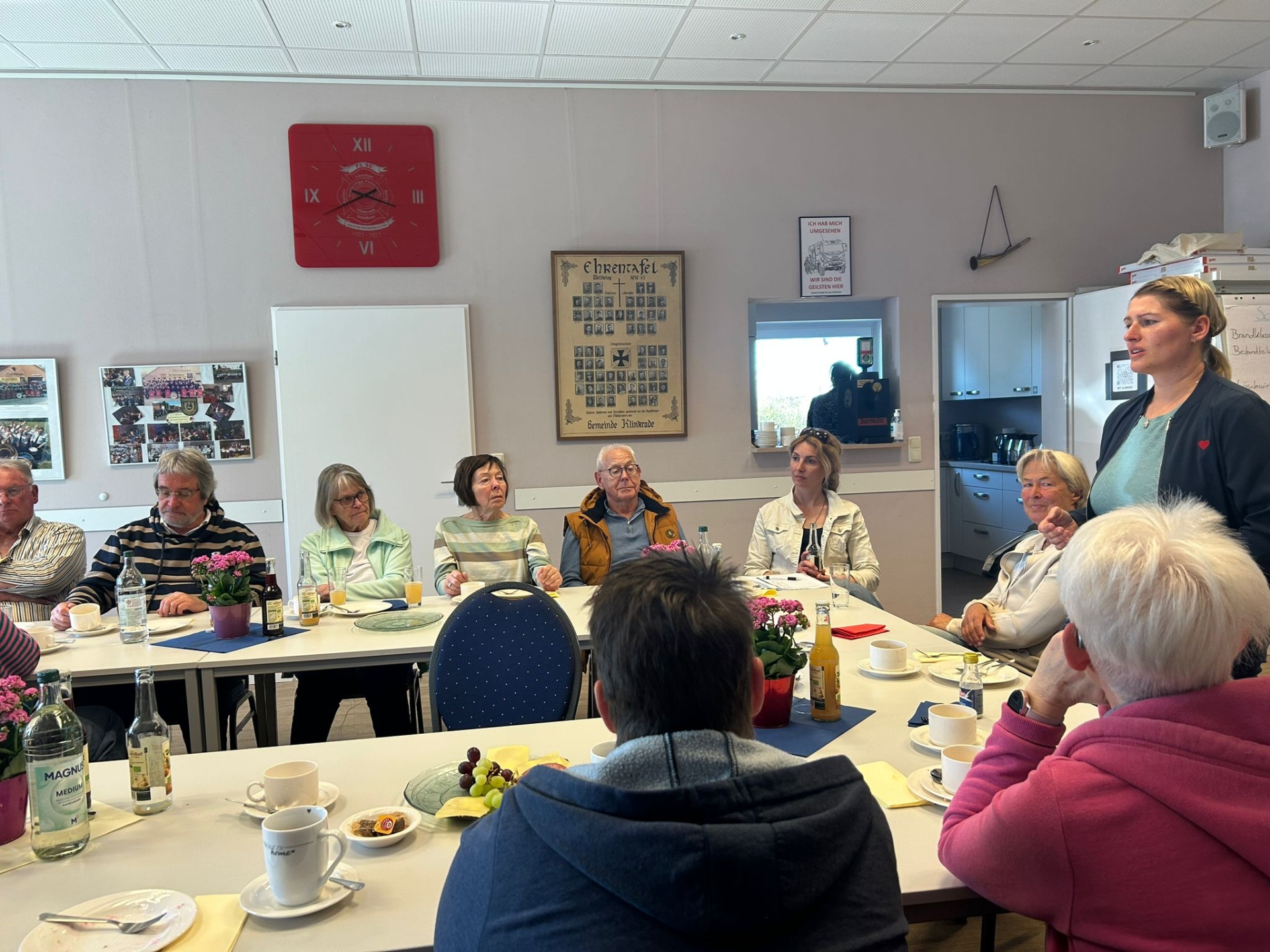 Mehrere Seniorinnen und Senioren sitzen an gedeckten Tischen in einem Gemeinschaftsraum und hören einer stehenden Frau zu, die spricht. Auf den Tischen stehen Kaffee, Getränke und kleine Speisen.