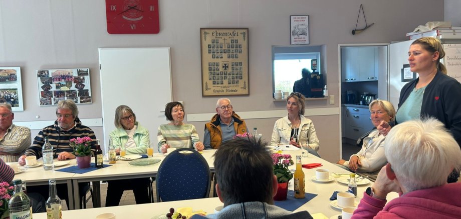 Mehrere Seniorinnen und Senioren sitzen an gedeckten Tischen in einem Gemeinschaftsraum und hören einer stehenden Frau zu, die spricht. Auf den Tischen stehen Kaffee, Getränke und kleine Speisen.