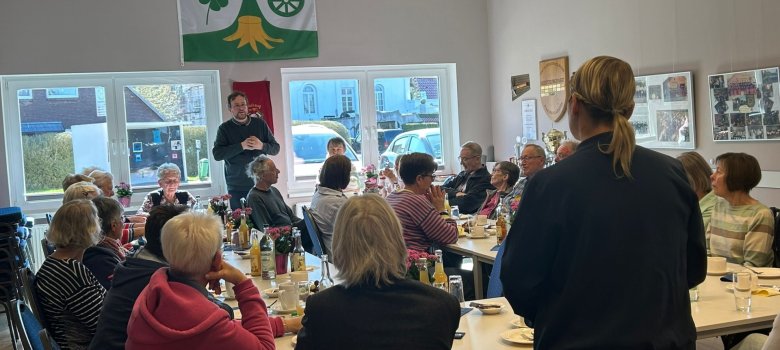 Ein voll besetzter Raum mit Seniorinnen und Senioren beim gemeinsamen Kaffeetrinken. Im Hintergrund spricht ein Mann vor der Gruppe, während die Anwesenden aufmerksam zuhören.