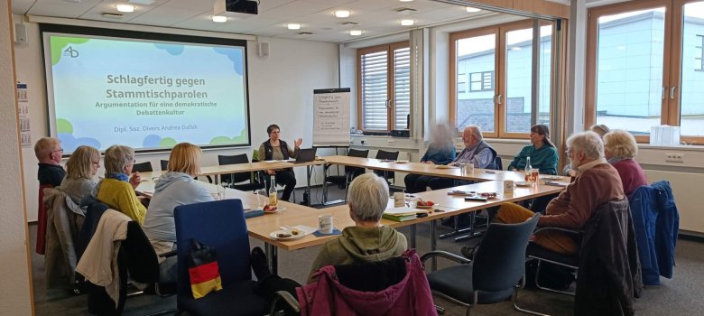 „In einem hellen Seminarraum sitzen mehrere Teilnehmende in U-Form an Tischen und hören einer Referentin zu. Auf der Leinwand ist das Thema ‚Schlagfertig gegen Stammtischparolen‘ zu sehen. Auf den Tischen stehen Getränke und kleine Speisen, die Atmosphäre ist aufmerksam und ruhig.