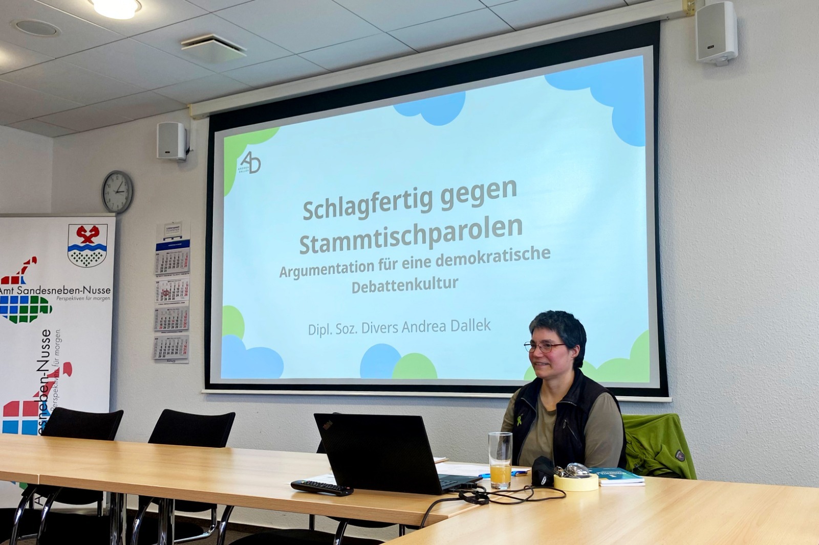 Eine Referentin sitzt an einem Tisch vor einer Leinwand mit der Präsentation ‚Schlagfertig gegen Stammtischparolen – Argumentation für eine demokratische Debattenkultur‘. Vor ihr stehen ein Laptop und ein Glas Saft. Der Raum wirkt wie ein Besprechungsraum im Amt Sandesneben-Nusse.