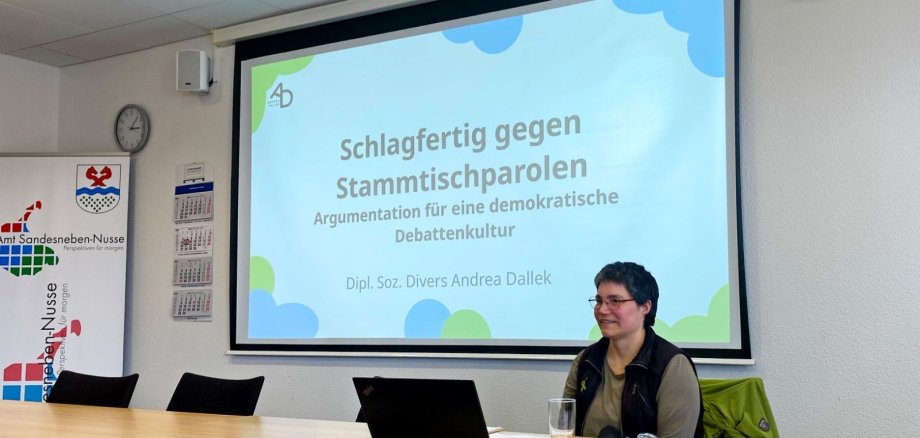 Eine Referentin sitzt an einem Tisch vor einer Leinwand mit der Präsentation ‚Schlagfertig gegen Stammtischparolen – Argumentation für eine demokratische Debattenkultur‘. Vor ihr stehen ein Laptop und ein Glas Saft. Der Raum wirkt wie ein Besprechungsraum im Amt Sandesneben-Nusse.