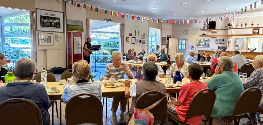 Zahlreiche Seniorinnen und Senioren sitzen gemeinsam an gedeckten Tischen in einem Saal. Im Hintergrund begleitet ein Musiker mit Gitarre die Veranstaltung.