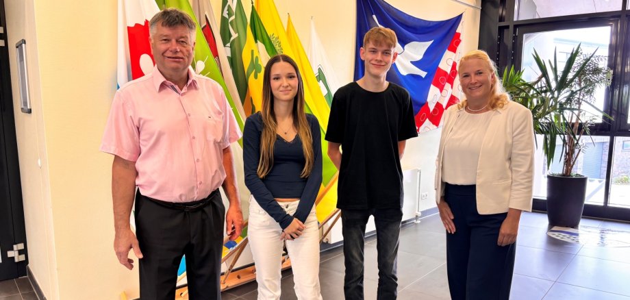 Amtsvorsteher Ulrich Hardtke und die leitende Verwaltungsbeamtin Tina Knuth begrüßen die neuen Auszubildenden Lina Hopke und Shawn Mesenbrink im Foyer des Amtes Sandesneben-Nusse. Die vier Personen stehen vor den Flaggen der amtsangehörigen Gemeinden und blicken in die Kamera.