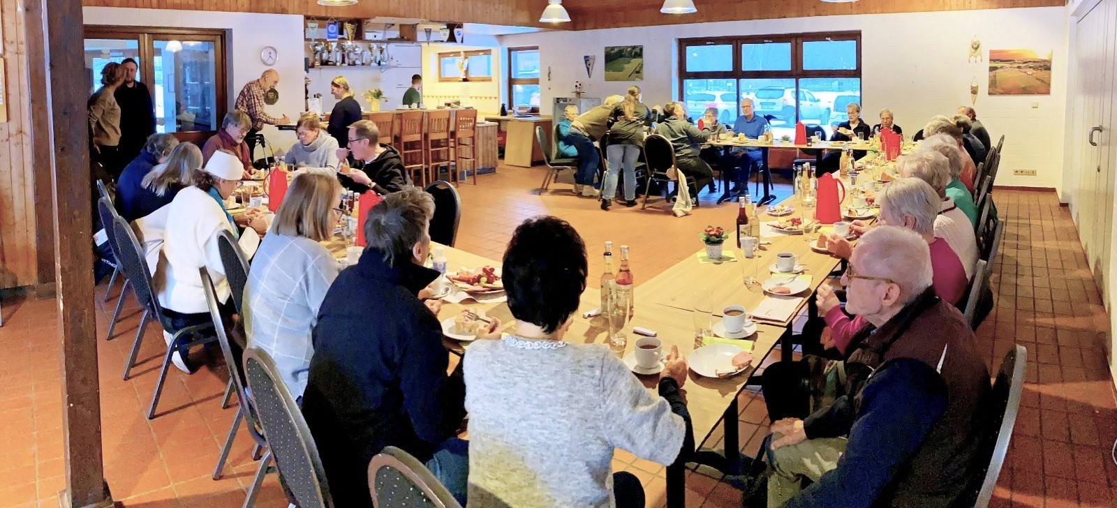 Mehrere Seniorinnen und Senioren sitzen an langen Tischen in einem Gemeindehaus und nehmen an einem gemeinsamen Kaffeetrinken teil.