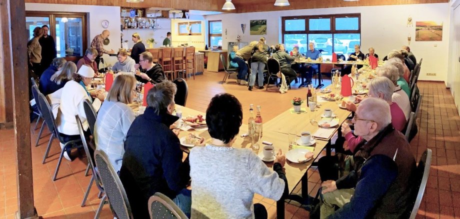 Mehrere Seniorinnen und Senioren sitzen an langen Tischen in einem Gemeindehaus und nehmen an einem gemeinsamen Kaffeetrinken teil.