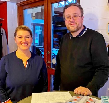 Zwei Personen stehen an einem Stehtisch in einem Innenraum. Vor ihnen liegen Unterlagen und Informationsmaterial.