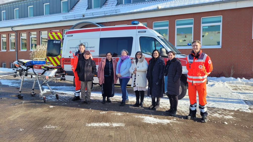 Mehrere Teilnehmende und zwei DRK-Mitarbeiter stehen vor einem Rettungswagen auf einem verschneiten Hof. Neben ihnen befindet sich eine Krankentrage mit Ausrüstung. Im Hintergrund ist ein Gebäude mit Backsteinfassade zu sehen.