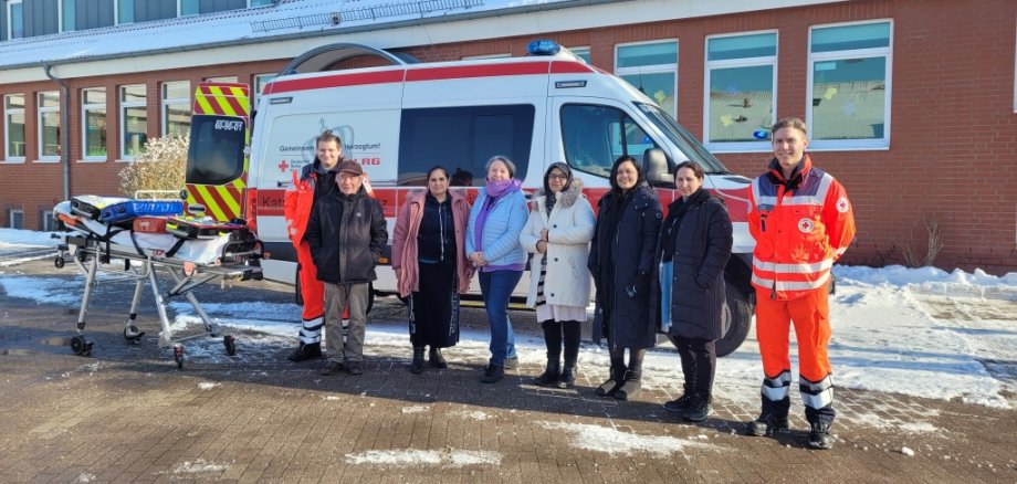 Mehrere Teilnehmende und zwei DRK-Mitarbeiter stehen vor einem Rettungswagen auf einem verschneiten Hof. Neben ihnen befindet sich eine Krankentrage mit Ausrüstung. Im Hintergrund ist ein Gebäude mit Backsteinfassade zu sehen.