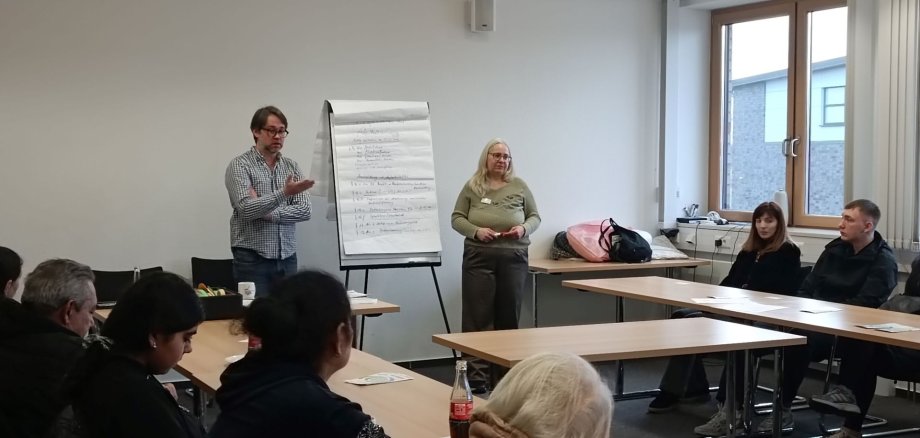 Zwei Personen stehen in einem Besprechungsraum vor einer Gruppe von Teilnehmenden. Ein Mann spricht und gestikuliert, neben ihm steht eine Frau. Hinter ihnen befindet sich ein Flipchart mit handschriftlichen Notizen. Die Teilnehmenden sitzen an Tischen in U-Form und hören zu.