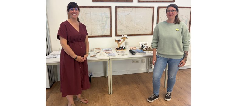 Zwei Frauen stehen neben einem Informationsstand mit Broschüren und kleinen Fläschchen. Im Hintergrund hängen historische Karten an der Wand. Die Aufnahme dokumentiert die Präsentation eines Angebots oder Projekts im Rahmen einer öffentlichen Veranstaltung.