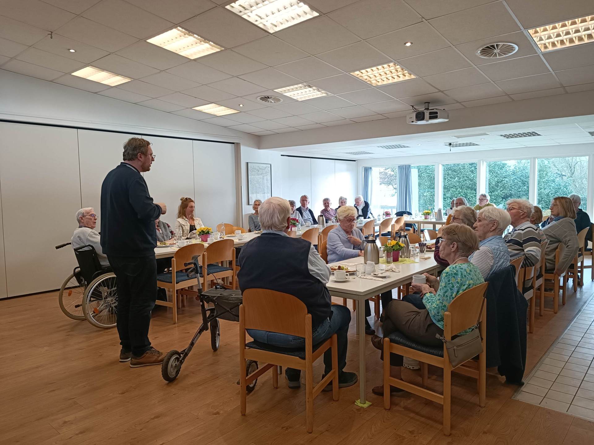 Mehrere ältere Menschen sitzen an Tischen in einem Gemeinschaftsraum, trinken Kaffee und unterhalten sich, während eine Person vorne spricht.