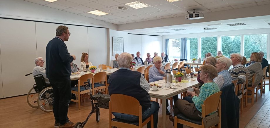 Mehrere ältere Menschen sitzen an Tischen in einem Gemeinschaftsraum, trinken Kaffee und unterhalten sich, während eine Person vorne spricht.