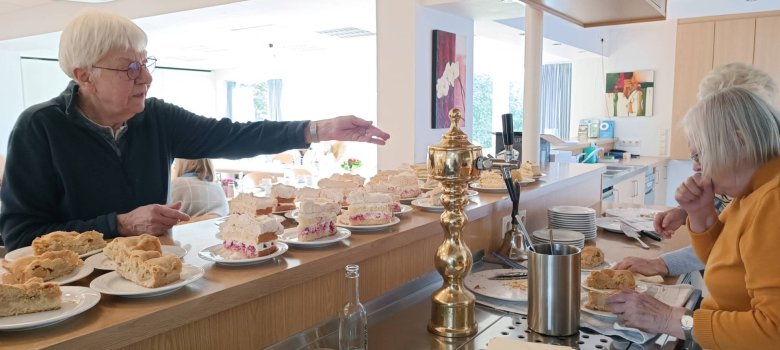 Zwei ältere Frauen richten an einer Theke mehrere Stücke Kuchen auf Tellern an, während im Hintergrund weitere Gäste sitzen.