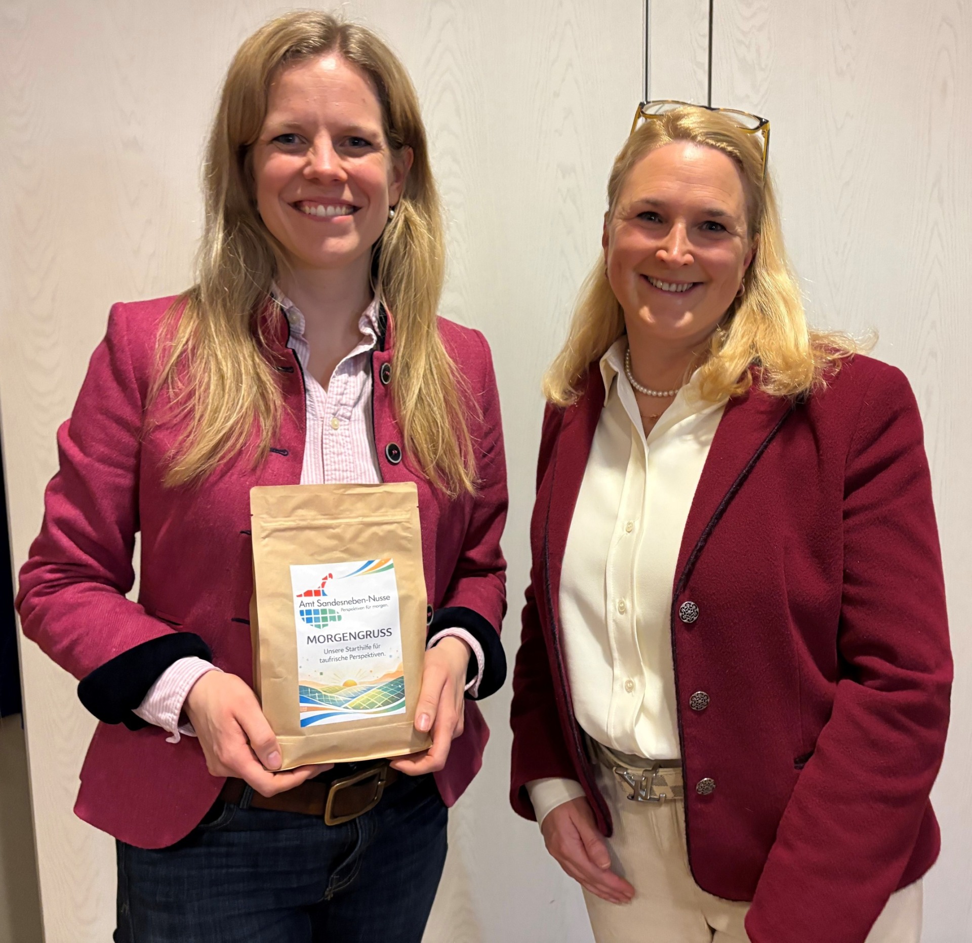 Cornelia Schmachtenberg, Landwirtschaftsministerin des Landes Schleswig-Holstein (links), und Tina Knuth, Leitende Verwaltungsbeamtin des Amtes Sandesneben-Nusse (rechts), stehen nebeneinander und lächeln in die Kamera. Cornelia Schmachtenberg hält eine Packung Kaffee mit der Aufschrift „Morgengruß – Unsere Stärke für ländliche Perspektiven“ und dem Logo des Amtes Sandesneben-Nusse in den Händen. Beide tragen bordeauxfarbene Blazer und stehen vor einer hellen Innenwand.