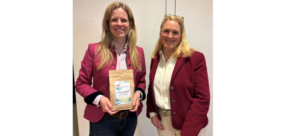 Cornelia Schmachtenberg, Landwirtschaftsministerin des Landes Schleswig-Holstein (links), und Tina Knuth, Leitende Verwaltungsbeamtin des Amtes Sandesneben-Nusse (rechts), stehen nebeneinander und lächeln in die Kamera. Cornelia Schmachtenberg hält eine Packung Kaffee mit der Aufschrift „Morgengruß – Unsere Stärke für ländliche Perspektiven“ und dem Logo des Amtes Sandesneben-Nusse in den Händen. Beide tragen bordeauxfarbene Blazer und stehen vor einer hellen Innenwand.