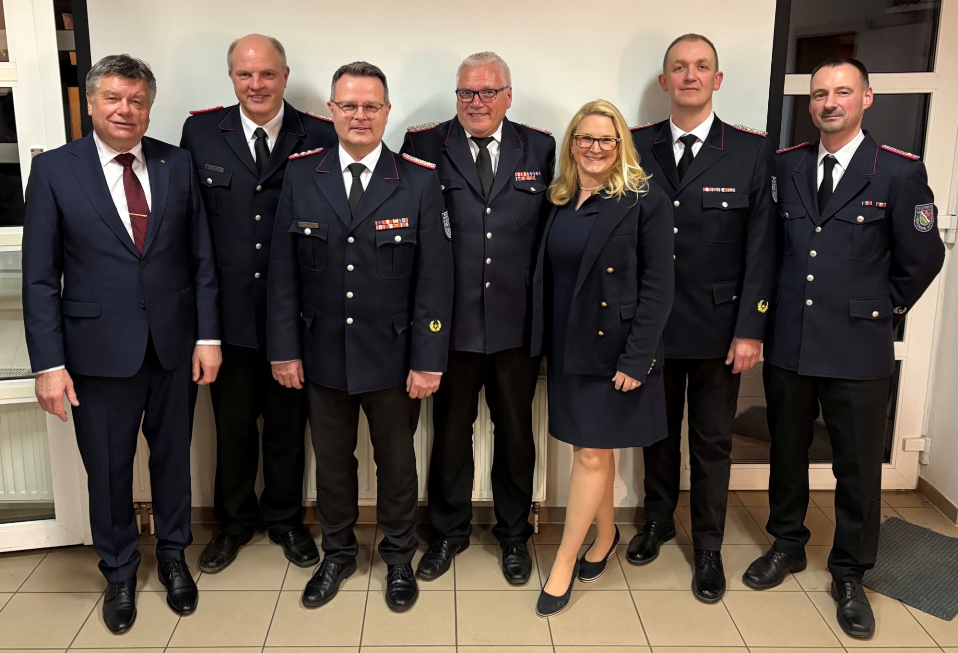 Sieben Personen stehen nebeneinander vor einer Leinwand in einem Innenraum. Sechs Männer tragen Feuerwehruniformen mit Abzeichen, eine Frau trägt ein dunkles Kostüm. Die Gruppe zeigt die aktuelle und ehemalige Amtswehrführung des Amtes Sandesneben-Nusse.